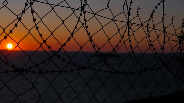 Orange sunset over ocean behind barbed wire fence. Barbed wire fence stands silhouetted against a vibrant orange sunset over a tranquil ocean, creating a poignant scene of confinement and beauty