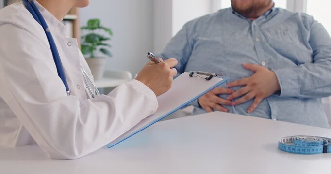 Doctor listens as a patient with obesity shows pain in the abdomen during a clinic consultation. Obesity patient reports stomach pain to physician sitting at desk in medical office.