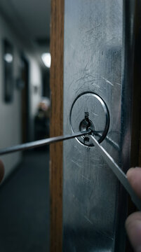 Hands of faceless burglar using lock picks to open door lock in dark office corridor. Professional locksmith working with picking tools. Crime concept with copy-space.