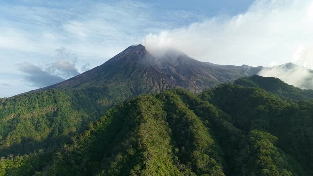 An aerial view of Mount Merapi surrounded by dense forest in Java, Indonesia. A view of volcanic terrain and hills under a clear blue sky.