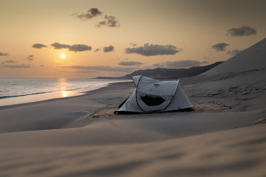 Socotra, Yemen - 05 April 2025: View of a white camping tent pitched on a sandy beach at sunset with rolling dunes and distant mountains.