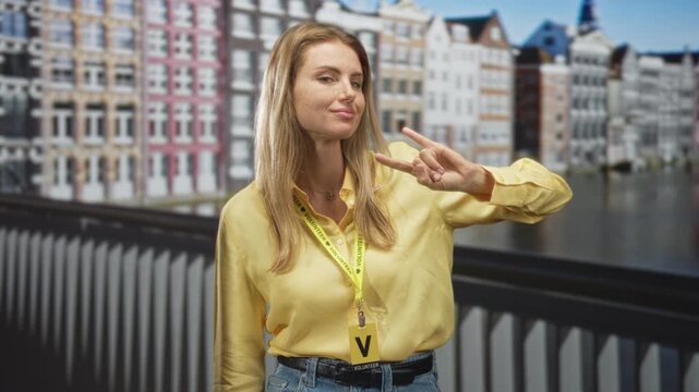 Blonde woman in yellow shirt wearing a volunteer lanyard and badge makes a peace sign with her hand while standing on a street bridge by canalfront row houses; playful confidence.