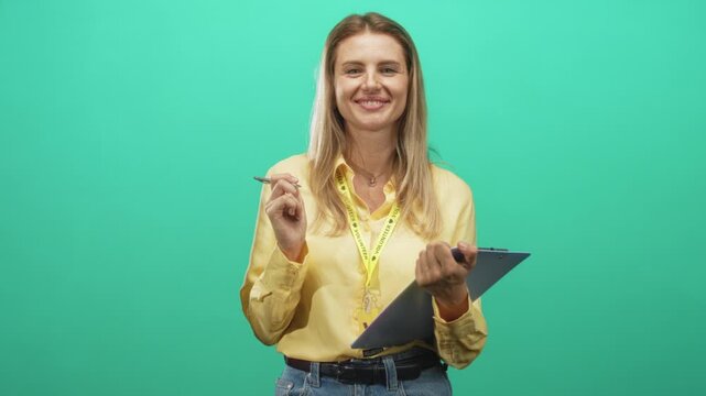 Blonde woman holding clipboard and pen, smiling while wearing a volunteer lanyard in studio with green backdrop; welcoming service.