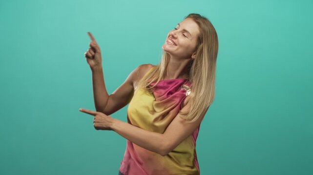 Woman pointing with index finger and hand toward upper left in studio teal backdrop, wearing colorful sleeveless top and smiling; playfulness.
