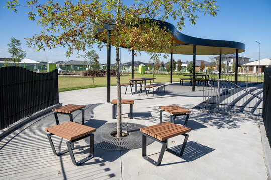 Contemporary public outdoor seating area in a newly developed suburban park in Australia, featuring seats arranged around a young shade tree.&nbsp;A modern shade shelter with picnic tables in background