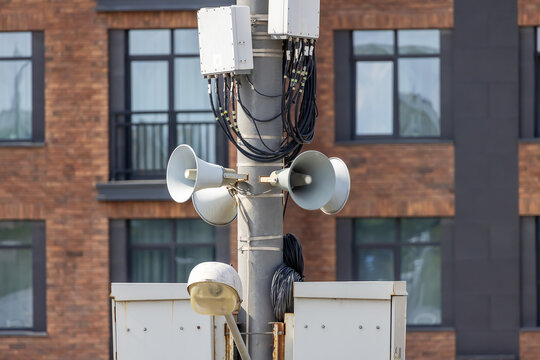 Public address system with multiple loudspeakers on metal pole against brick building. Outdoor siren and alert equipment for emergency notifications in city.
