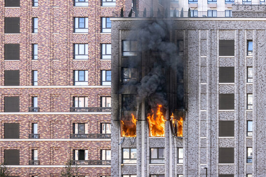 Dangerous fire incident in multi-story residential building with flames and black smoke from windows. Emergency situation with burning apartment in modern urban district.