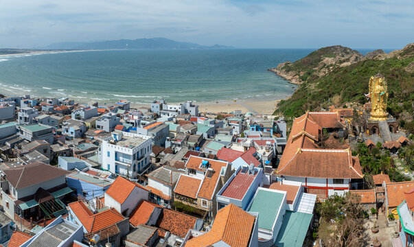 A panoramic view of the golden Buddha statue overlooking the coastal town of Nhon Ly, Quy Nhon, Binh Dinh, Vietnam, with wind turbines on the hills in the background.