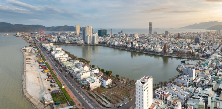 Aerial view of Sinh Thai Lake, Quy Nhon, Binh Dinh, Vietnam, showing the port with shipping containers and trucks, and the city skyline with modern buildings and mountains in the background.