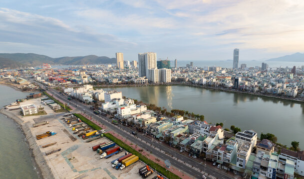 Aerial view of Sinh Thai Lake, Quy Nhon, Binh Dinh, Vietnam, showing the port with shipping containers and trucks, and the city skyline with modern buildings and mountains in the background.