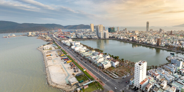 Aerial view of Sinh Thai Lake, Quy Nhon, Binh Dinh, Vietnam, showing the port with shipping containers and trucks, and the city skyline with modern buildings and mountains in the background.
