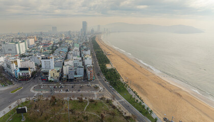 Obraz premium An aerial view of the Beach in Quy Nhon, Binh Dinh, Vietnam, shows a wide sandy shore with scattered beachgoers and the city skyline in the background under a cloudy sky.
