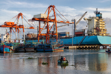 Naklejka premium A large container ship is loaded at a busy port in Quy Nhon, Binh Dinh, Vietnam, with smaller fishing boats docked nearby. Two people in a small boat navigate the water.