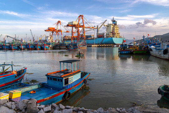 A busy port in Quy Nhon, Binh Dinh, Vietnam, with fishing boats and a large cargo ship being loaded by cranes. The scene captures the daily maritime activity.