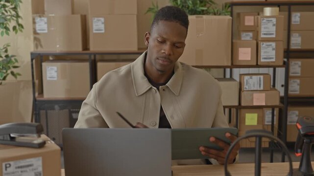 Man holds tablet and writes with pen while checking a laptop amid stacked parcels in a building; logistics concentration.