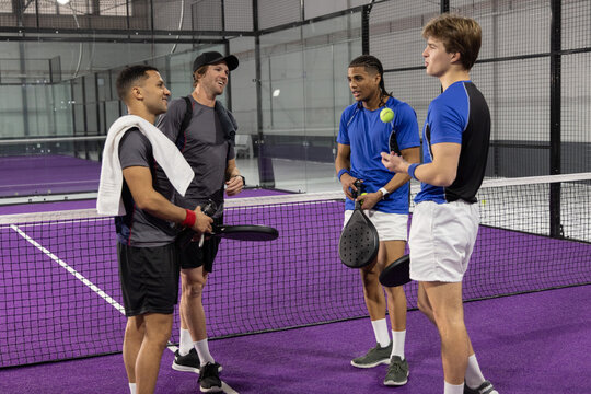 Diverse male teammates standing and talking at net on purple court with padel paddles, yellow ball