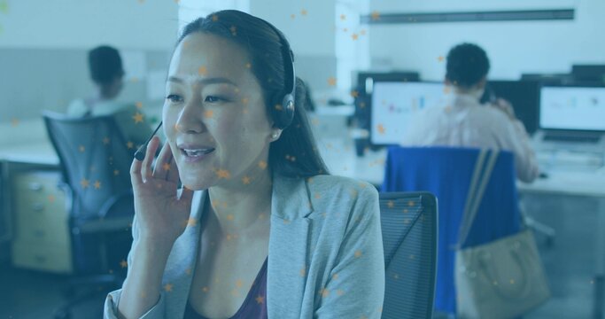 Speaking woman in grey blazer wearing headset handling calls in open-plan office, with monitors