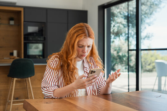 Young redhead woman sitting at a kitchen table looking frustrated and confused while reading her mobile phone, reacting to a message or unexpected problem with technology, service, or bill.