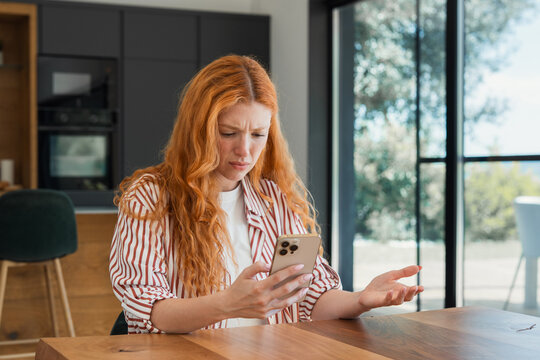 Red-haired woman frustrated and confused by smartphone at kitchen table reacting to app error