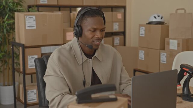 Man with headset smiles while typing on laptop and handling parcels at desk in building; cheerful small business.
