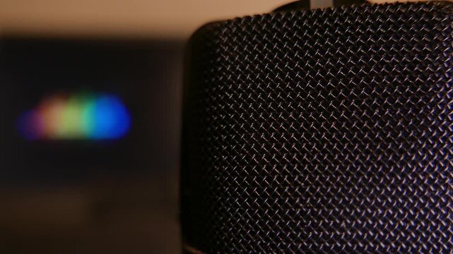Slow zoom out revealing details of a professional microphone in a recording studio. Blurred colorful sound levels moving intensely on a laptop screen in the background. Close-up: a podcast microphone