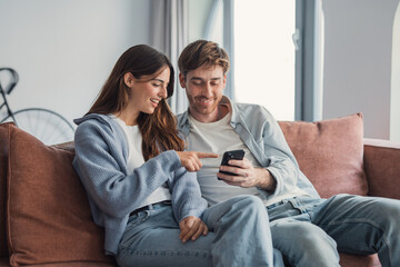 Obraz premium Young couple sitting on a sofa at home smiling and sharing a smartphone while relaxed together