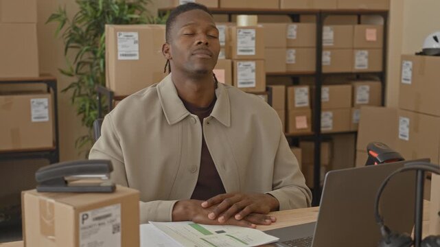 Man with hands on desk and head tilted back, eyes closed, seated at laptop amid stacked parcels with shipping labels visible in a building; fatigue logistics delay.