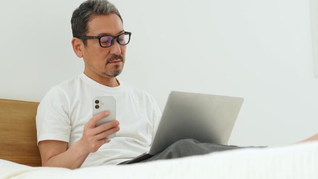 ベッドの上でスマートフォンとパソコンを使う中高年の男性