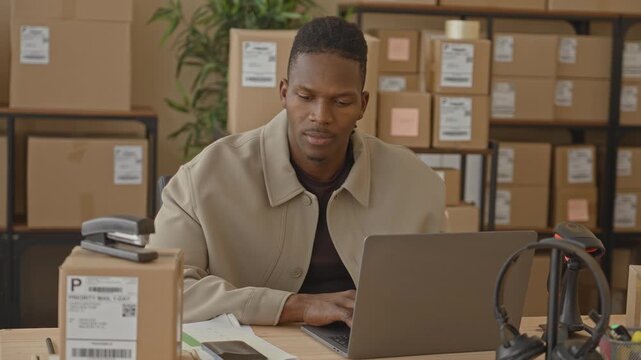 Man holds smartphone to ear and checks laptop among stacked parcels and barcode scanner on desk in a building; small business success joy.