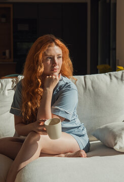 A redhead woman in comfortable pajamas sits on a light sofa holding a mug, bathed in warm morning sunlight, appearing calm and thoughtful while relaxing at home.