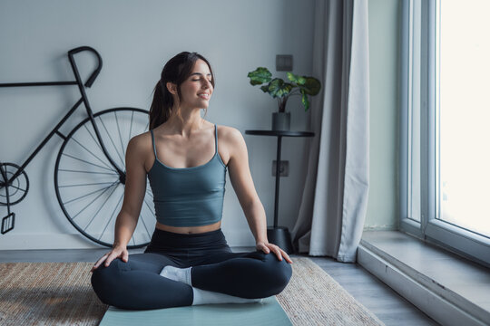 A smiling young woman sits cross-legged on a yoga mat, practicing meditation and breathing exercises in a bright living room. Calm, relaxed and focused, she enjoys a peaceful home fitness moment.