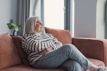 Obraz premium Elderly woman with gray hair and glasses sits on a comfortable sofa near a bright window, looking thoughtful and relaxed in her living room, enjoying a quiet moment at home.