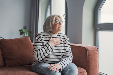 Obraz premium An elderly woman sits on a soft couch, hand to her chest, gazing out a bright window with a thoughtful, reflective expression in a calm, cozy living room setting.