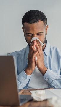 Tired young man blows his nose into a tissue at a home desk while using a laptop, conveying cold symptoms, fatigue and remote work. Illness, allergy and self-care concepts in an indoor setting.