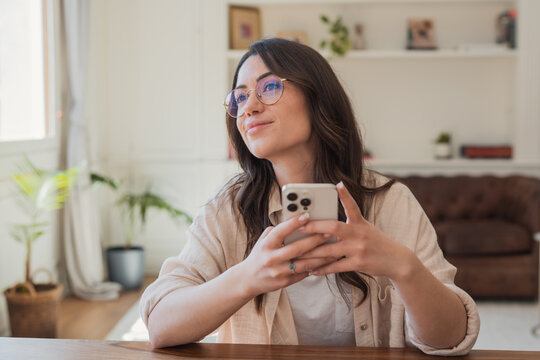 Young woman smiling and holding a smartphone at home while thoughtfully looking away
