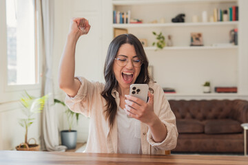 A young woman in glasses cheers with her fist while reading good news on her smartphone at home, showing joy, surprise and celebration in a cozy living room environment.