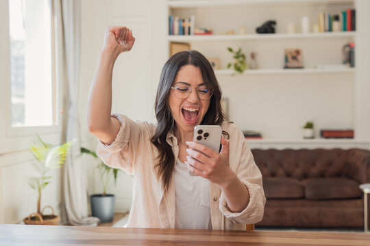 A young woman in glasses cheers with her fist while reading good news on her smartphone at home, showing joy, surprise and celebration in a cozy living room environment.