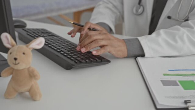 Man doctor in lab coat with stethoscope typing on computer keyboard, hands on keys, eyewear and tie visible in clinic building; calm professionalism.