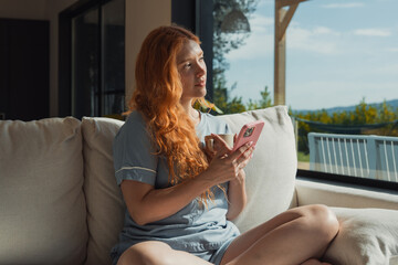 Obraz premium Redheaded woman sits on a couch holding a smartphone and coffee mug, gazing out a large window toward a balcony and scenic view, enjoying a relaxed, sunny morning at home.