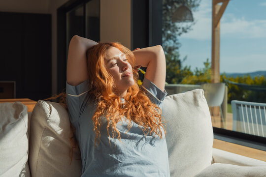 Red-haired woman lounges on a sofa with hands behind her head, basking in warm sunlight through large windows and enjoying a calm, peaceful moment of relaxation at home.