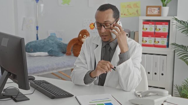 Man doctor holds phone to ear while writing with pen, stethoscope visible on desk in medical building; calm focus.