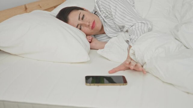 Young woman reaches hand for smartphone on mattress in a bedroom while lying under white duvet and pillow beside her; sleepy solitude.