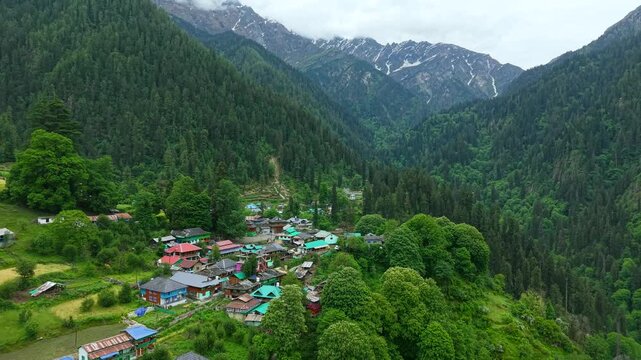 Grahan village with clustered colorful rooftops surrounded by dense pine forests in Parvati Valley, Himachal Pradesh, India. Aerial dolly backward pullout.