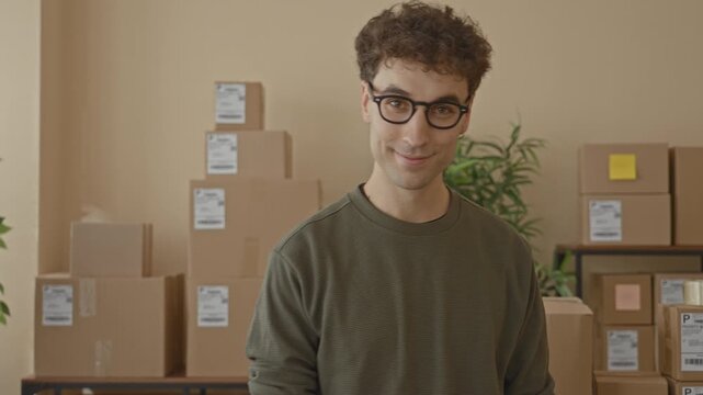 Young man smiles at camera amid stacked shipping boxes and parcels on shelves in a building; friendly service.