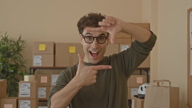 Man with packages frames his face with hands and points at camera while smiling amid stacked parcels on shelves inside a building; excitement.