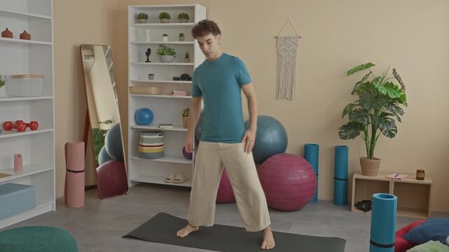 Man performing side stretch on yoga mat in studio, arm raised and hand on hip, surrounded by exercise balls and mirror; wellness serenity.
