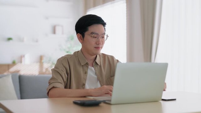 Happy young Asian man working on laptop in living room. Freelance worker male sitting at desk writing notes while watching webinar, studying online