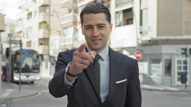 Man in suit points finger to camera on a busy city street beside a bus and storefronts, smiling with tie, lapel and watch visible; confidence invitation.