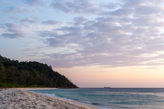 Koh Adang Beach at sunset with calm ocean waves. Lush green hills in the background. Sandy shoreline in Thailand.