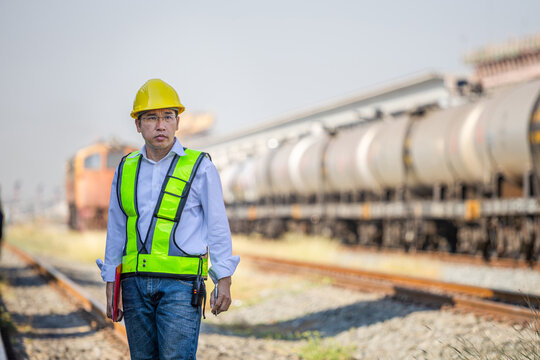 Asian engineer wearing safety helmet and vest standing on railway track with oil tank train background, Inspector holding clipboard and walkie-talkie standing near freight train in industrial station